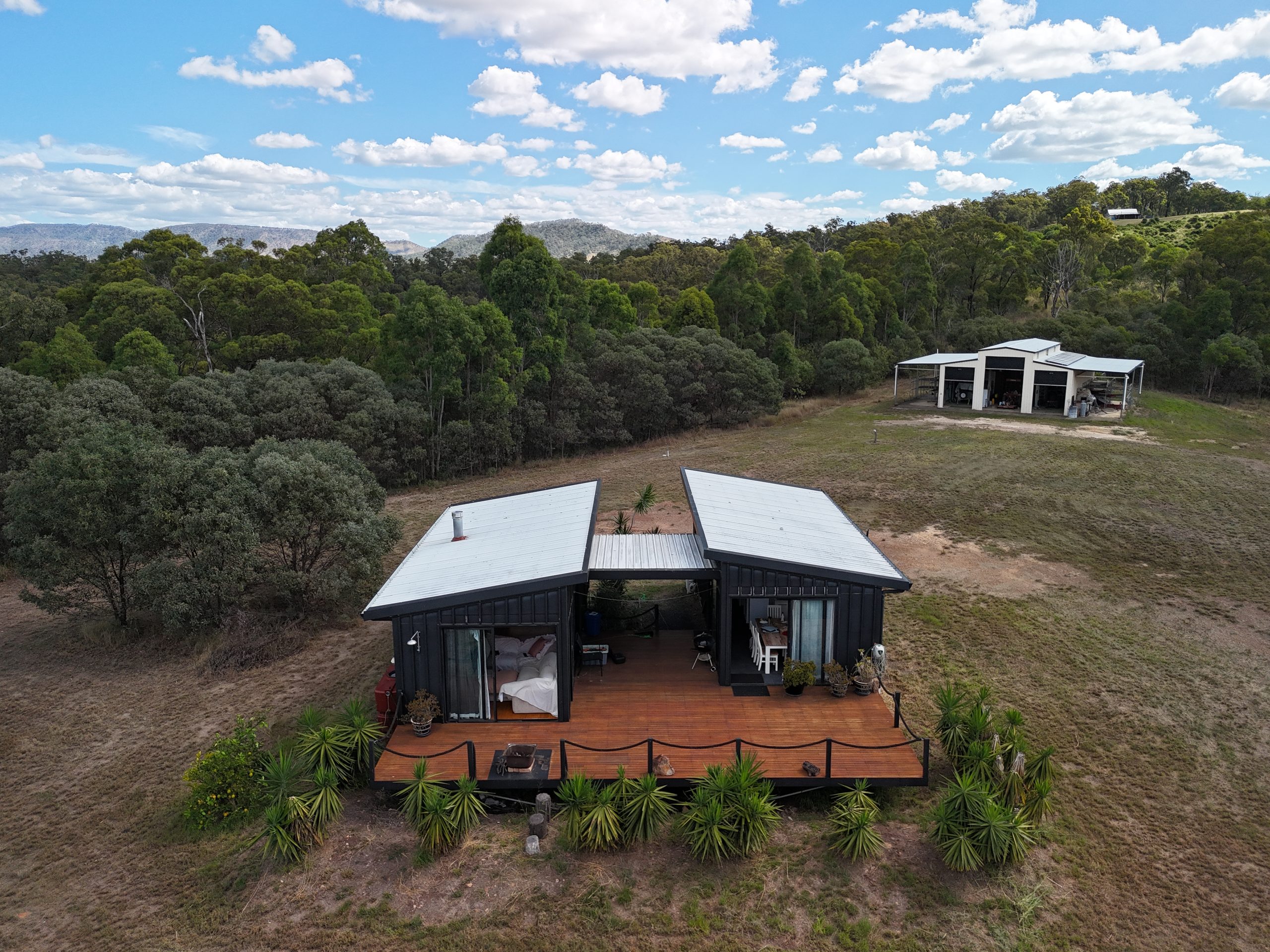 Golden-hour aerial view of a modern cabin with a timber deck and a separate machinery shed, set on acreage land with mountain views in the background.