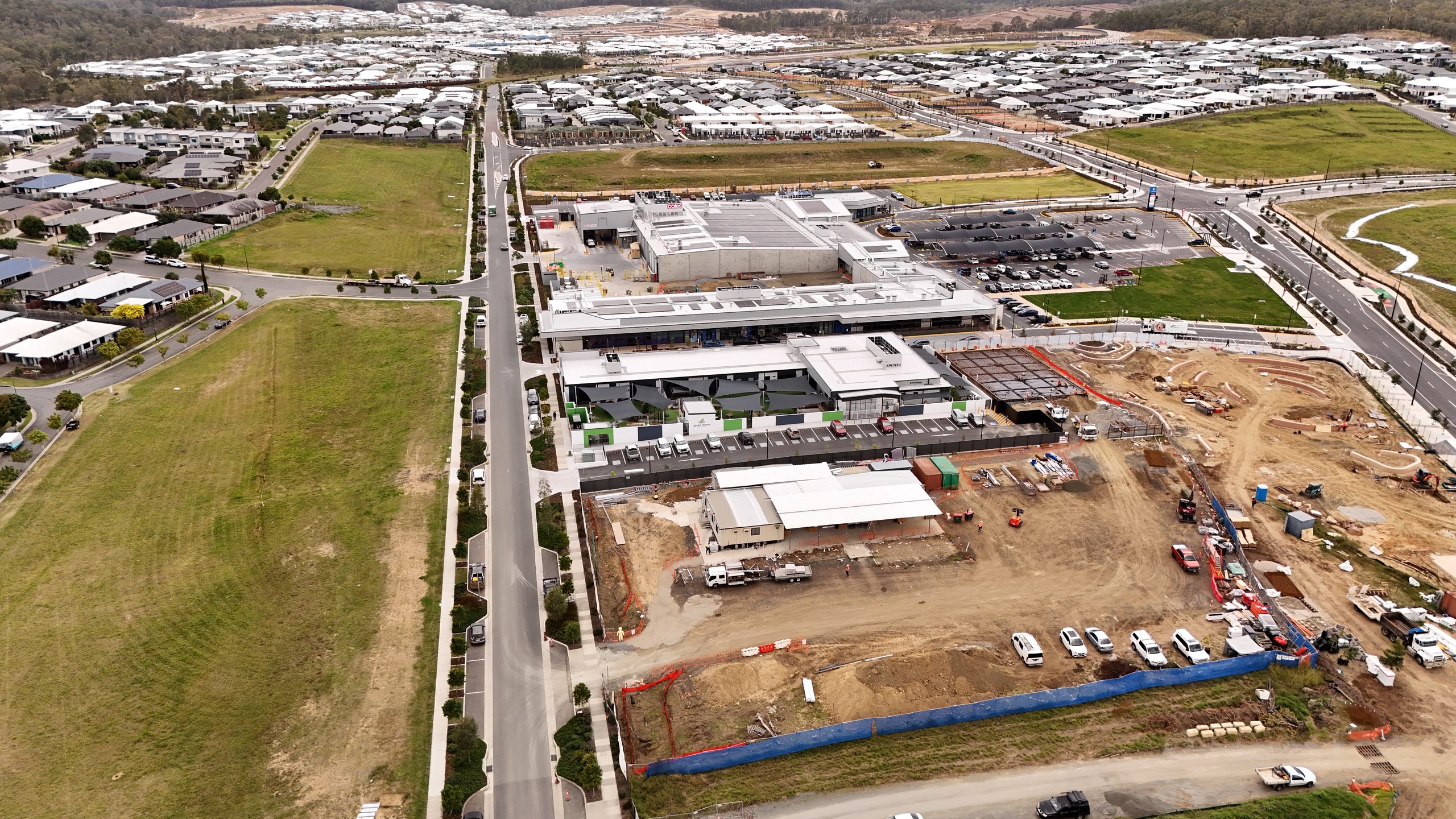 Aerial view of a commercial shopping precinct under construction in Brisbane, showing completed retail buildings, earthworks, parked machinery and site vehicles alongside a new residential subdivision.