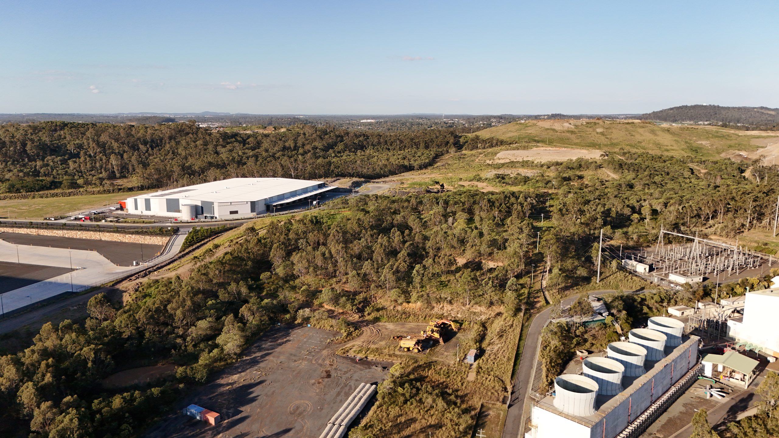 Wide aerial view of an industrial warehouse and electrical substation set among bushland, with cleared earthworks and distant hills under a partly cloudy sky.