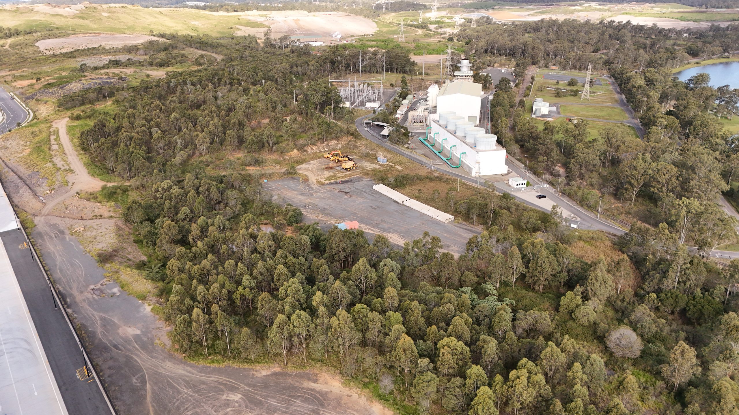 Aerial view of an industrial power facility under development, showing white processing buildings, an electrical substation, excavation equipment and forested surrounds.