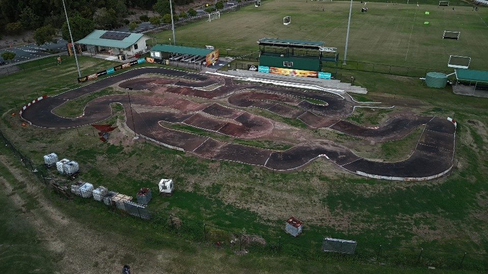 Aerial view of a winding dirt motocross track with grandstands and sponsor banners at a recreation facility, alongside sports fields at dusk.
