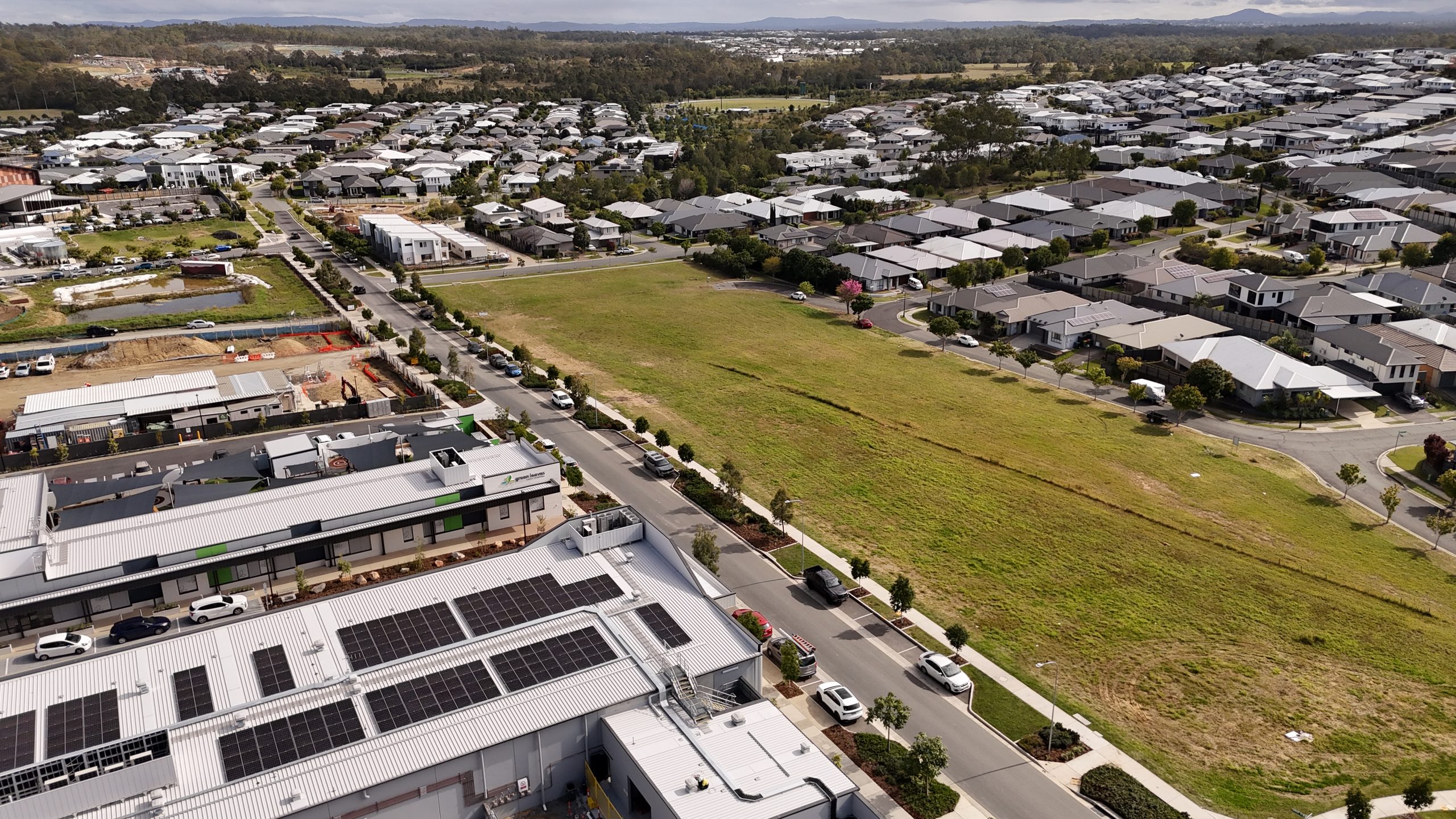 Aerial overview of a new housing estate with rows of rooftops, streets and a vacant grassed lot under a dramatic cloudy sky.