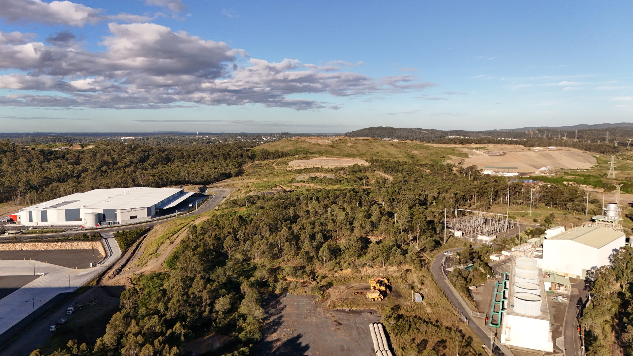 Aerial view of an electrical substation and white industrial warehouse building surrounded by bushland, with transmission towers visible in the distance.