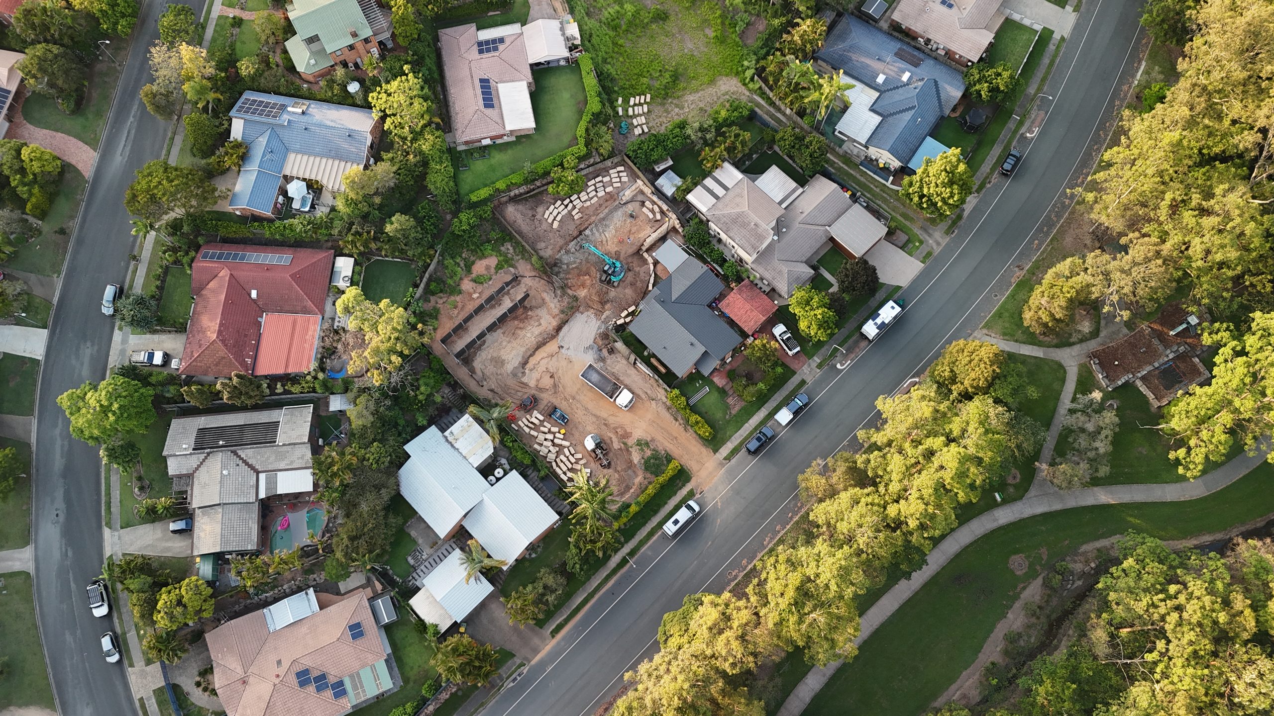 Aerial view of a house under construction on an infill block in a Brisbane suburb, with an excavator, building materials and earthworks visible between established homes.