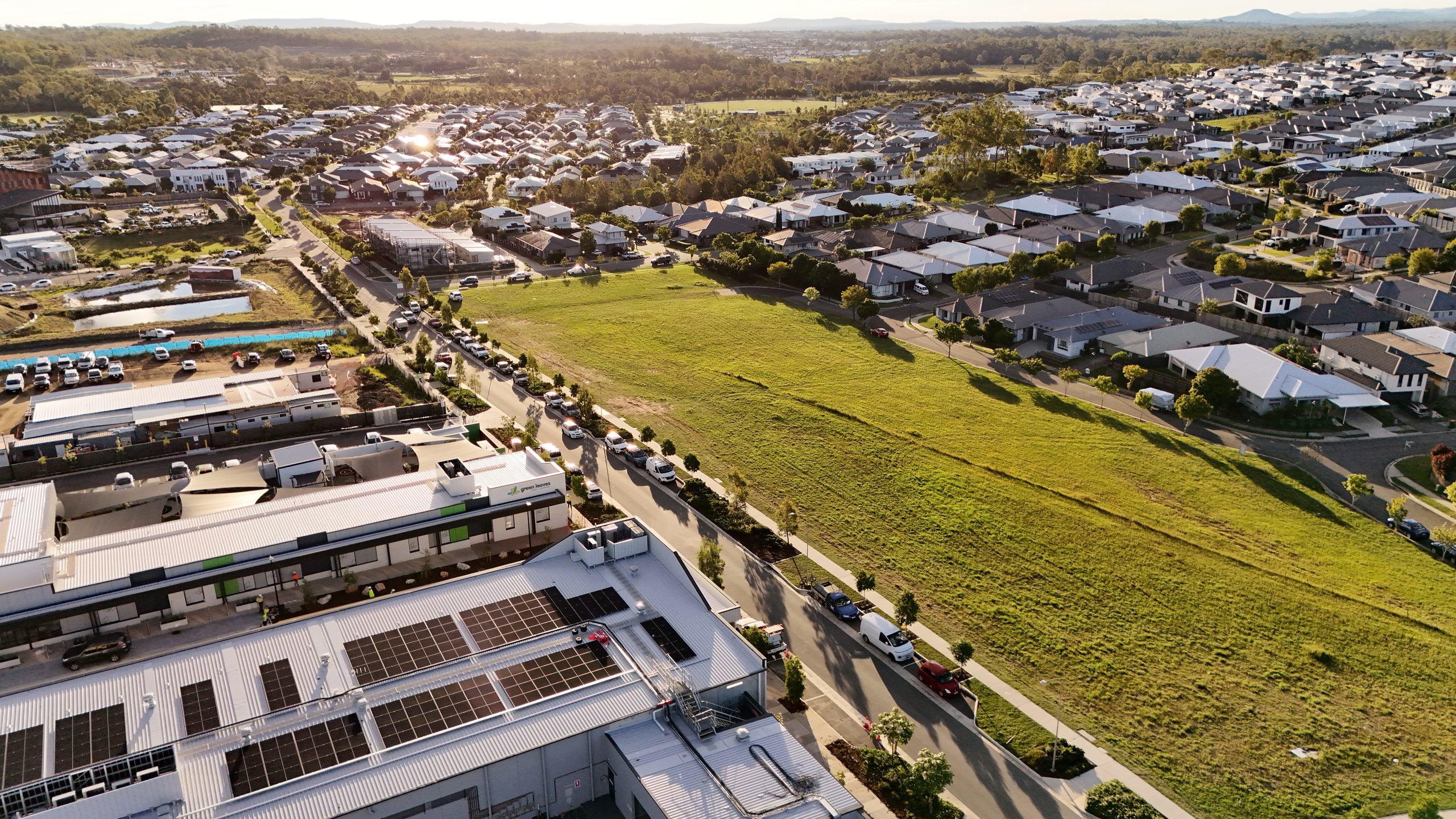 Aerial view of a residential land development at sunrise, showing established homes, a vacant grassed lot and a shopping centre under construction.