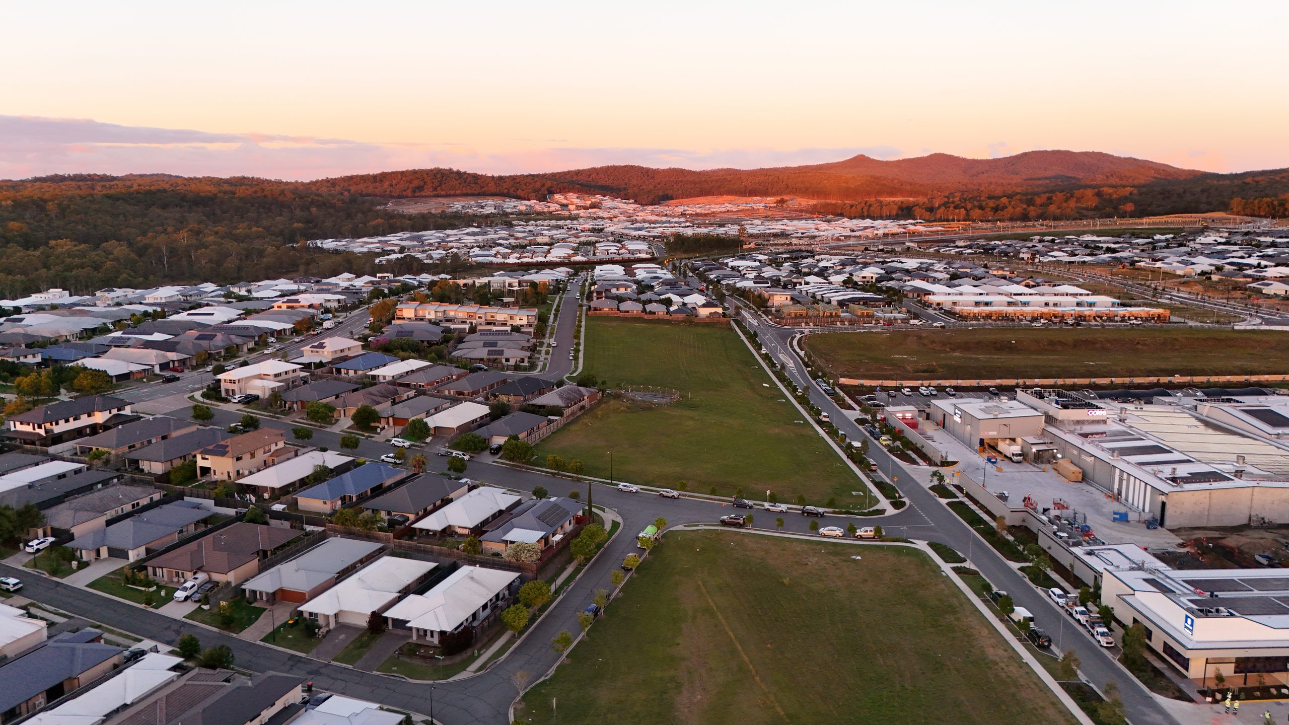 Wide aerial panorama at sunset over a Brisbane residential subdivision, showing rooftops, streets and green space with hills silhouetted on the horizon.