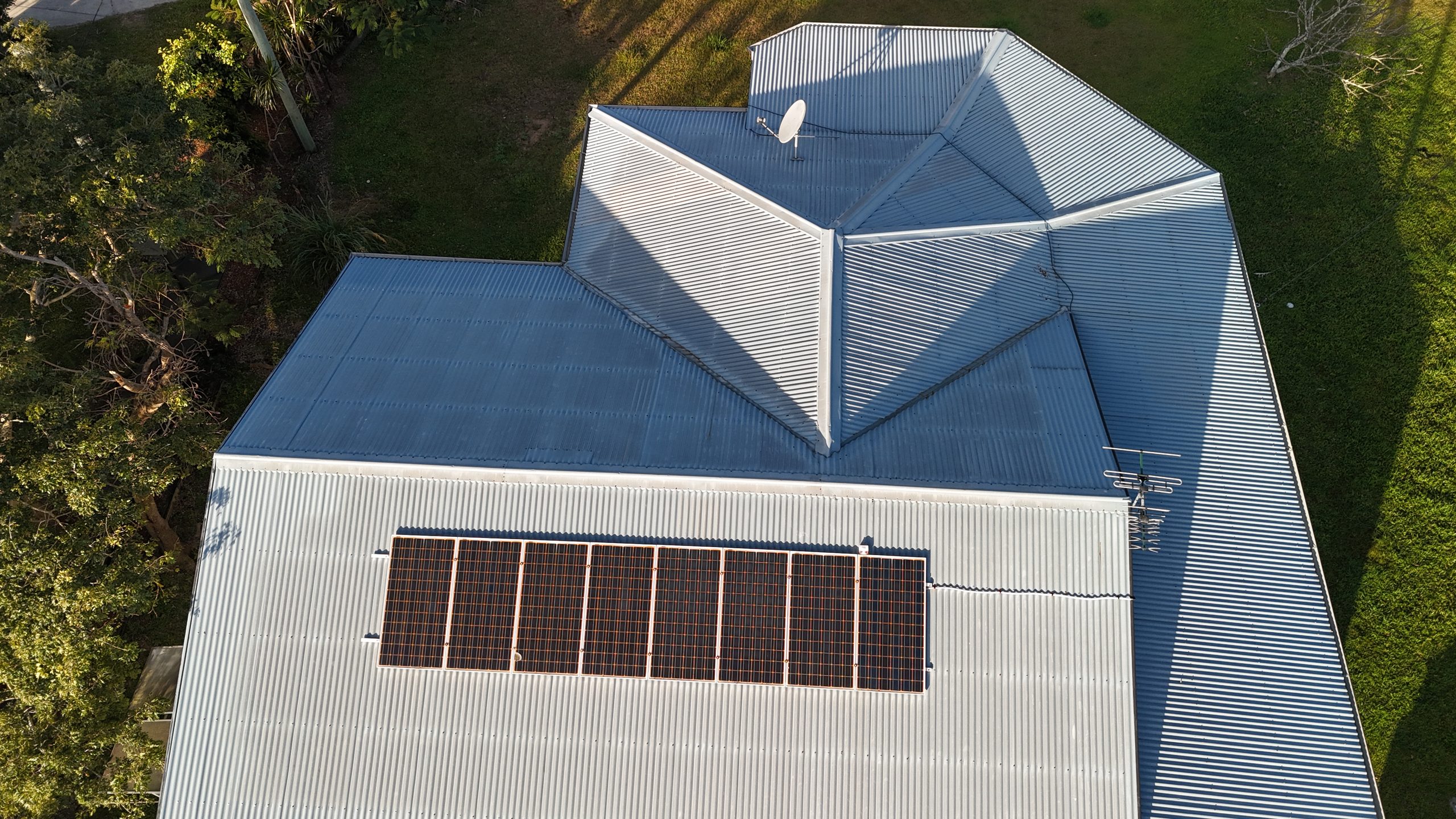 Aerial view of a metal-roofed shed with a rooftop solar panel array, satellite dish and antenna, surrounded by lawn and trees.