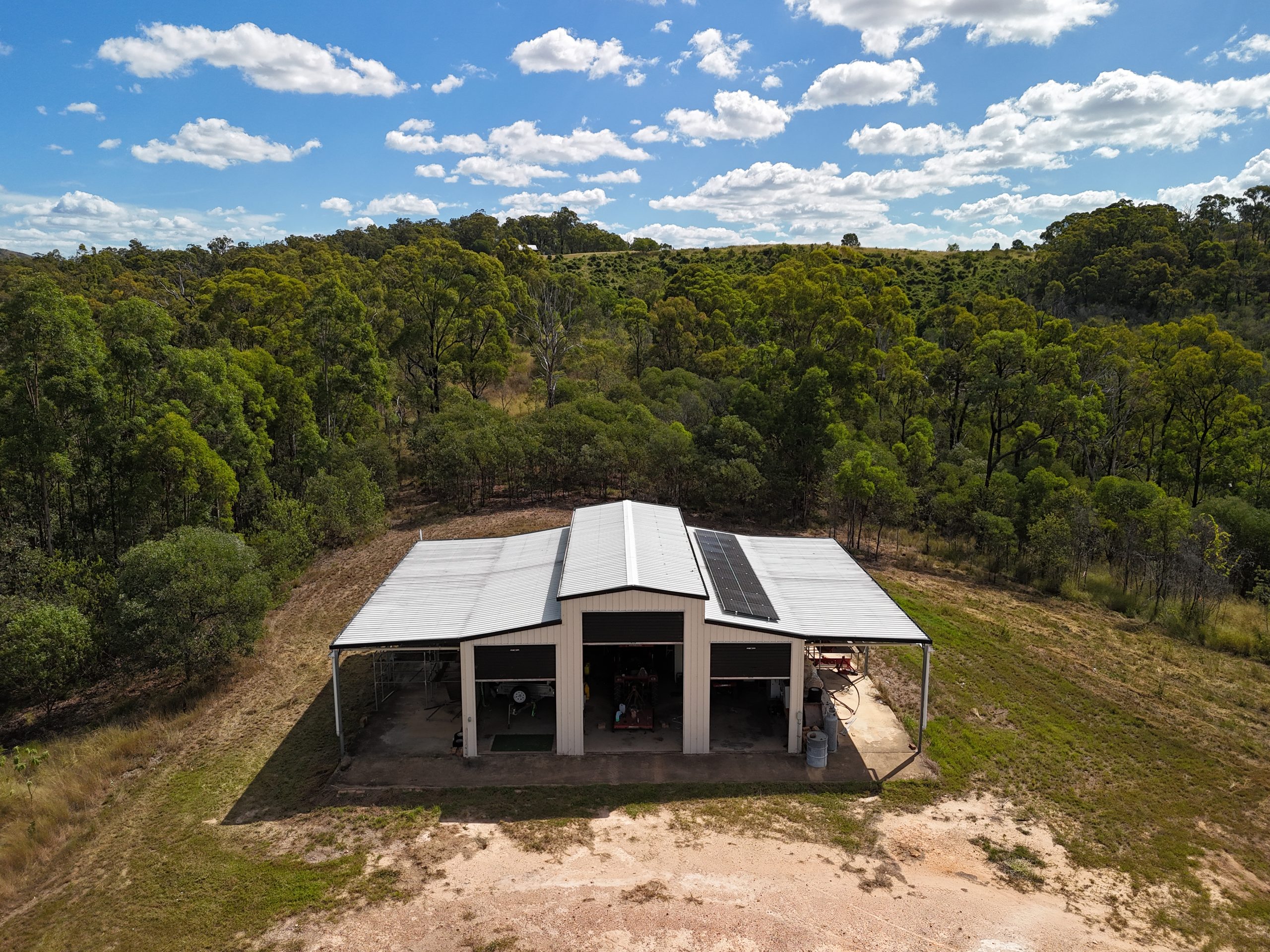 Aerial view of a large rural shed with a rooftop solar panel array, set on acreage land with forested hills and a modern cabin nearby.