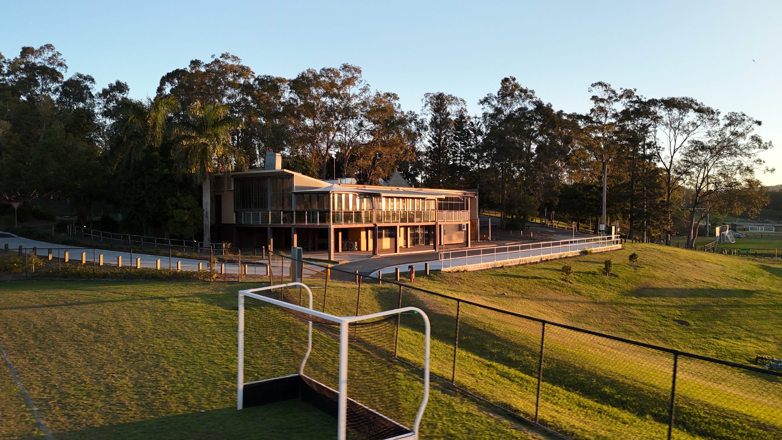 Golden-hour aerial view of a two-storey sports clubhouse with wraparound balconies, framed by trees and a soccer field in the foreground.