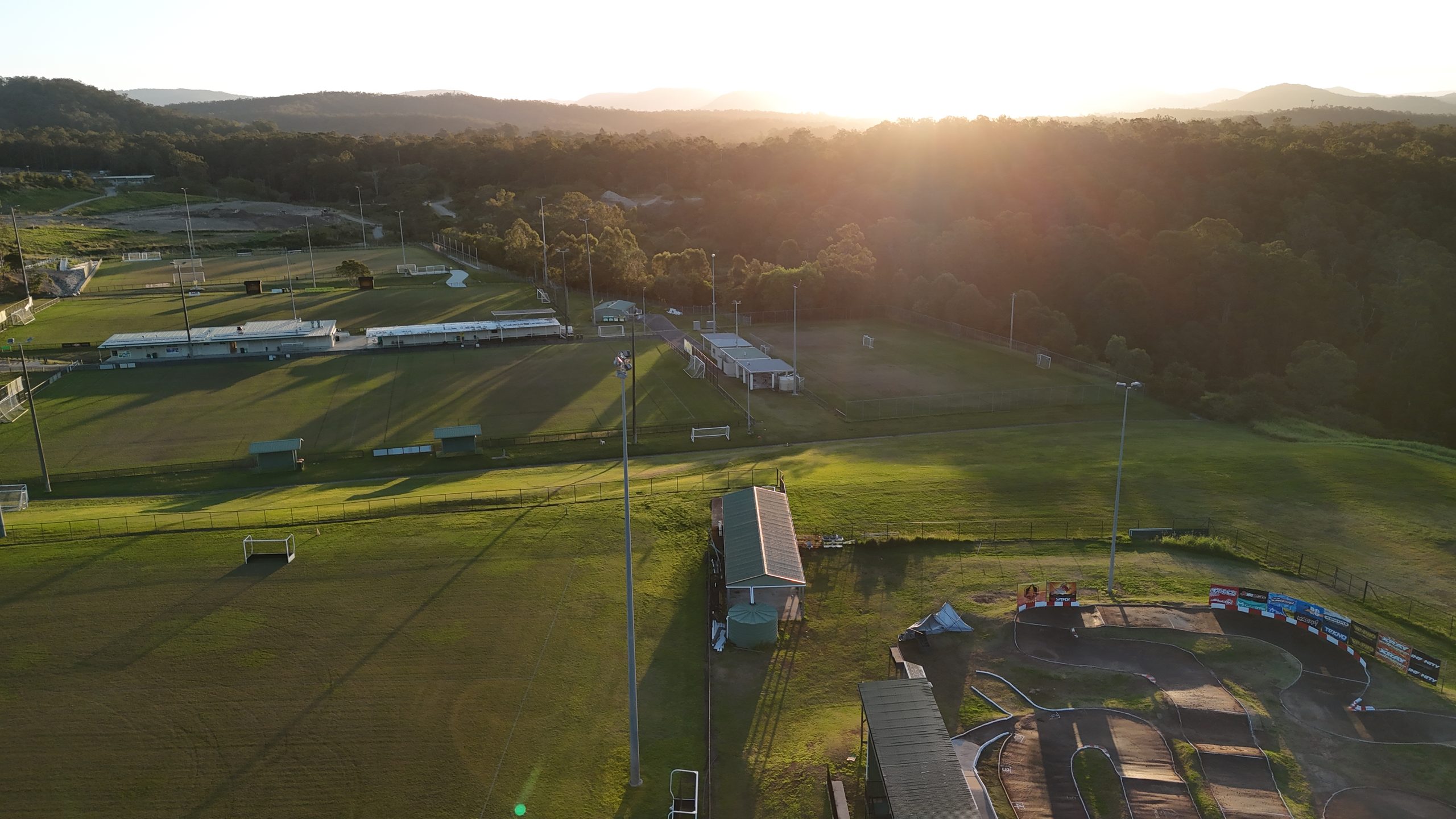 Aerial view at sunset of a multi-field sports complex with soccer pitches, a clubhouse and a recreation track, backlit by warm evening light.