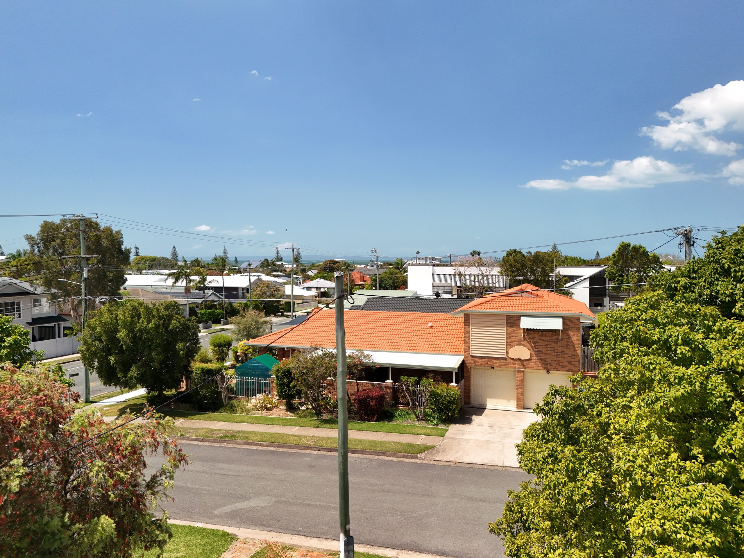 Aerial view of a single-storey brick home with an orange and black tiled roof on a suburban street in Brisbane, under a clear blue sky.