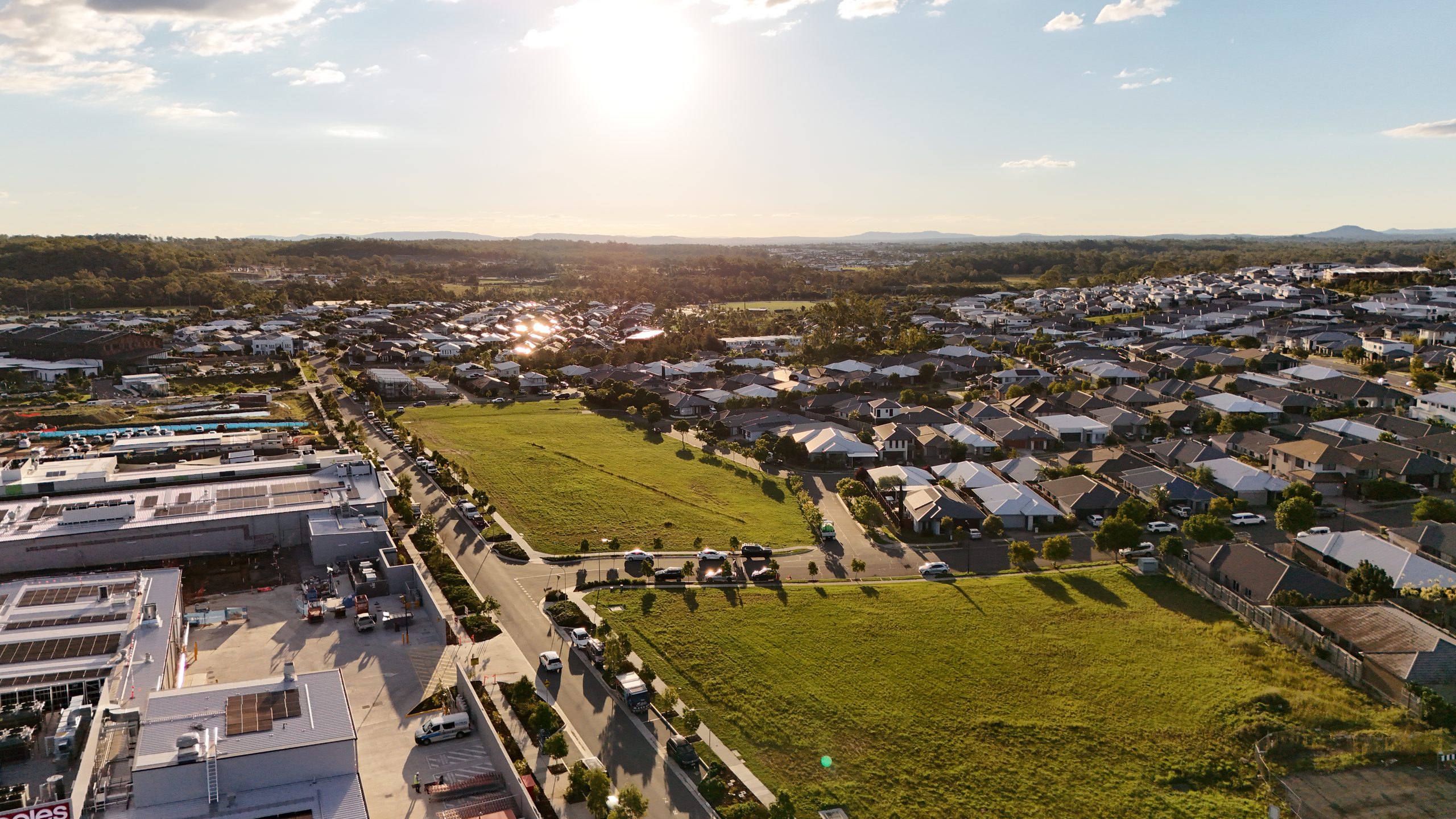 Aerial view at sunset of vacant subdivided land bordered by new housing and a shopping centre, with warm light over distant hills.