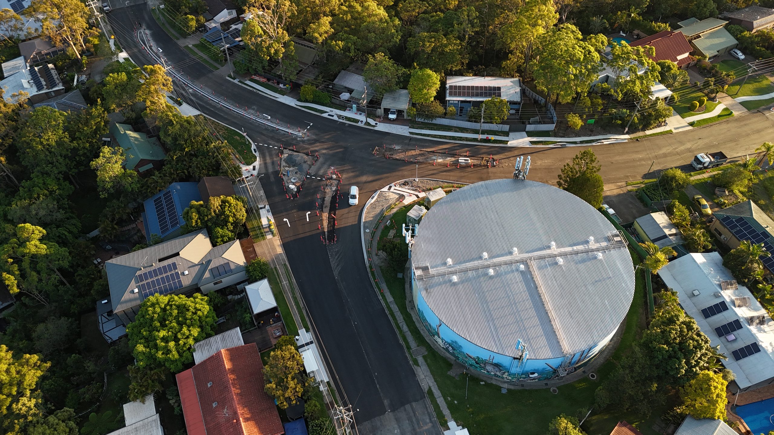 Top-down drone view of a circular water tower with a mural, surrounded by suburban road works, traffic cones and construction markings at golden hour.